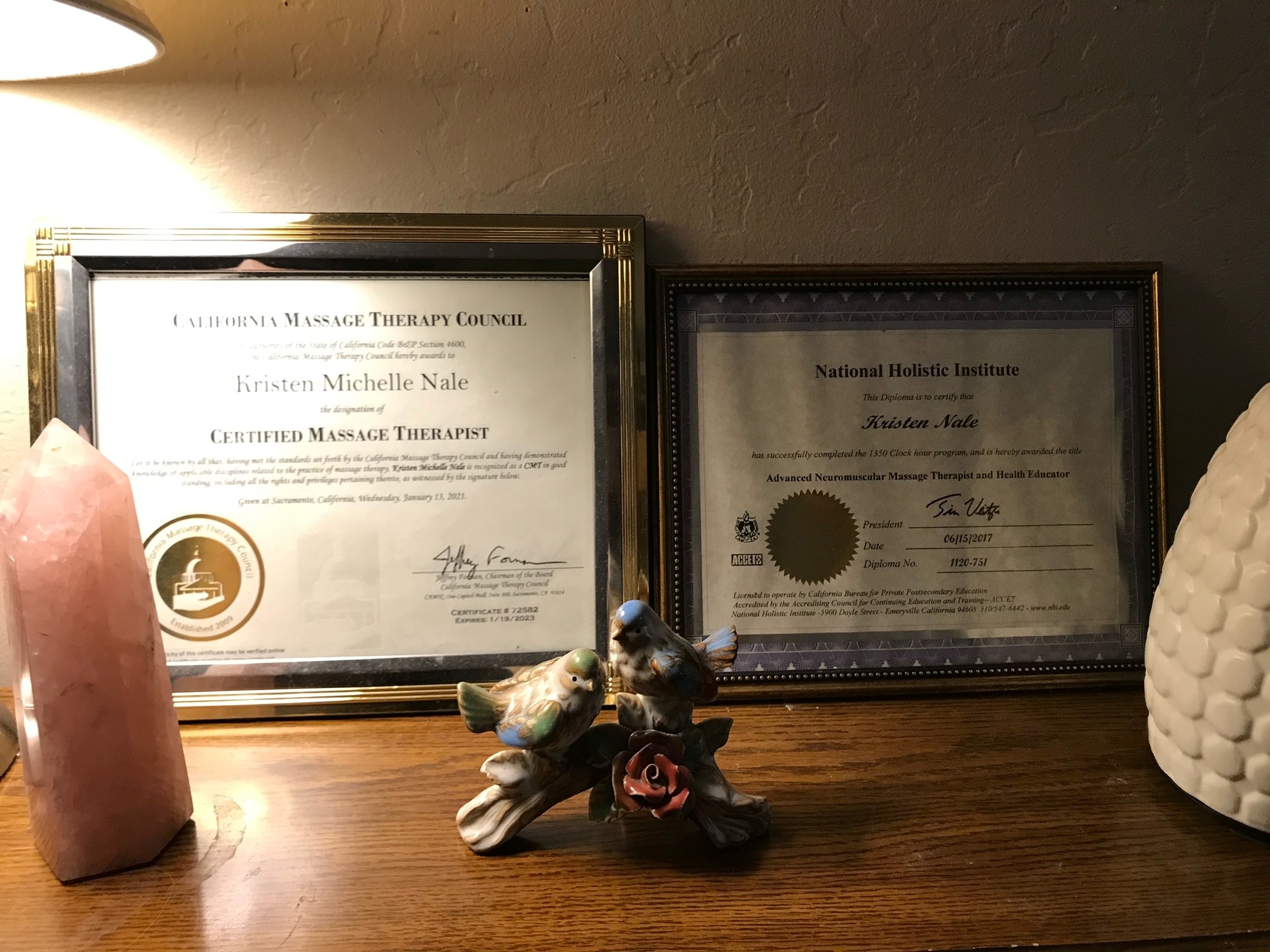 Two framed massage-therapy certificates issued to Kristen Michelle Nale, displayed next to a tall rose-quartz crystal and a ceramic bird figurine