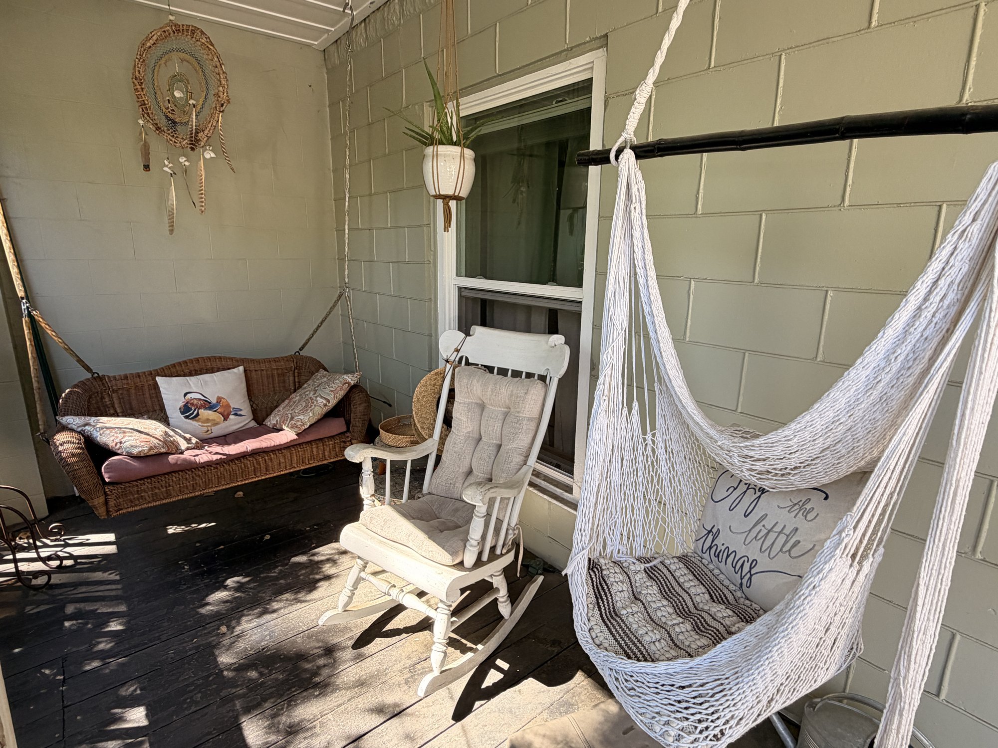 Front porch with a white wicker porch swing, a wooden rocking chair, and a knotted-rope hammock chair. A dreamcatcher on the wall and a hanging plant.