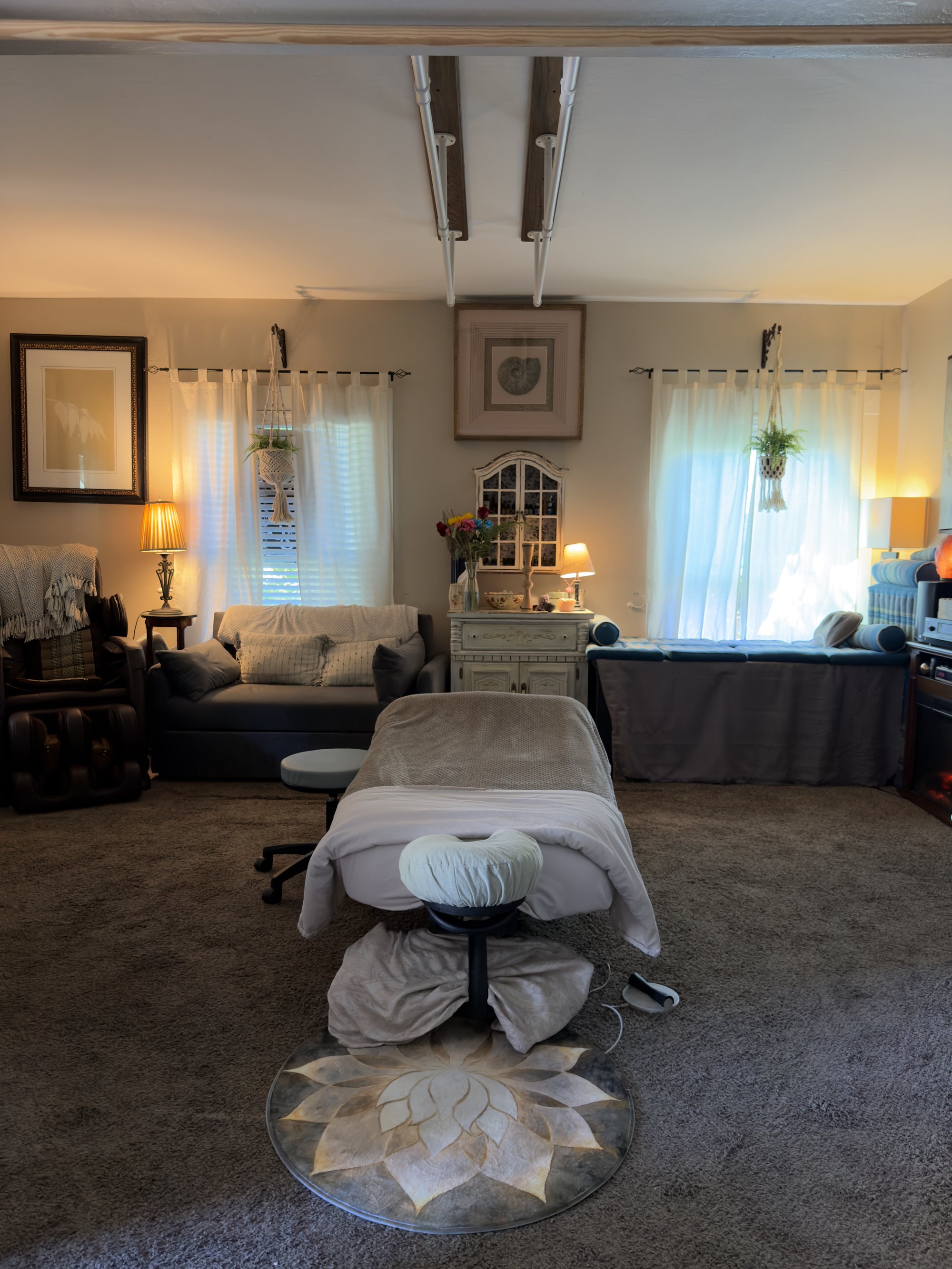 Wide view of the treatment room — massage table centered with white linens, two sheer-curtained windows, an antique cabinet between them, framed art, a grey day-bed sofa, and a small lotus mat under the table.